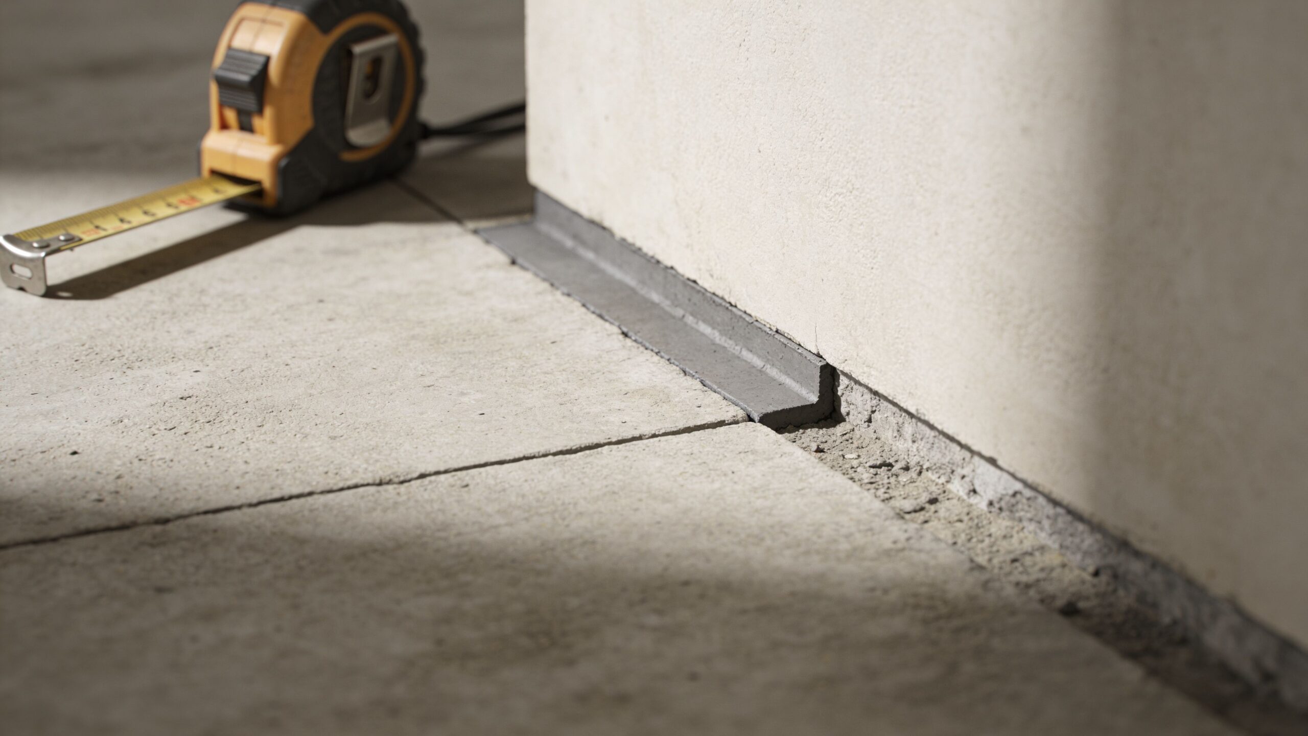 Et nærbillede af et betongulv under opbygning med en målebånd og kantbånd mod en væg.