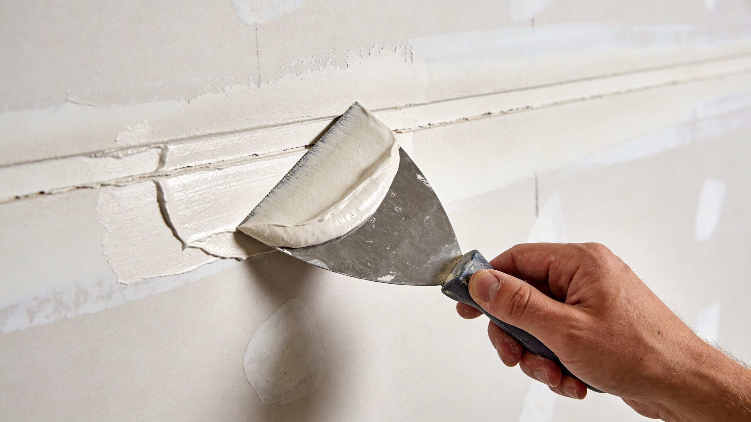 En hånd bruger en spartel til at påføre gipsspartelmasse på en samling mellem to gipsplader på væggen.