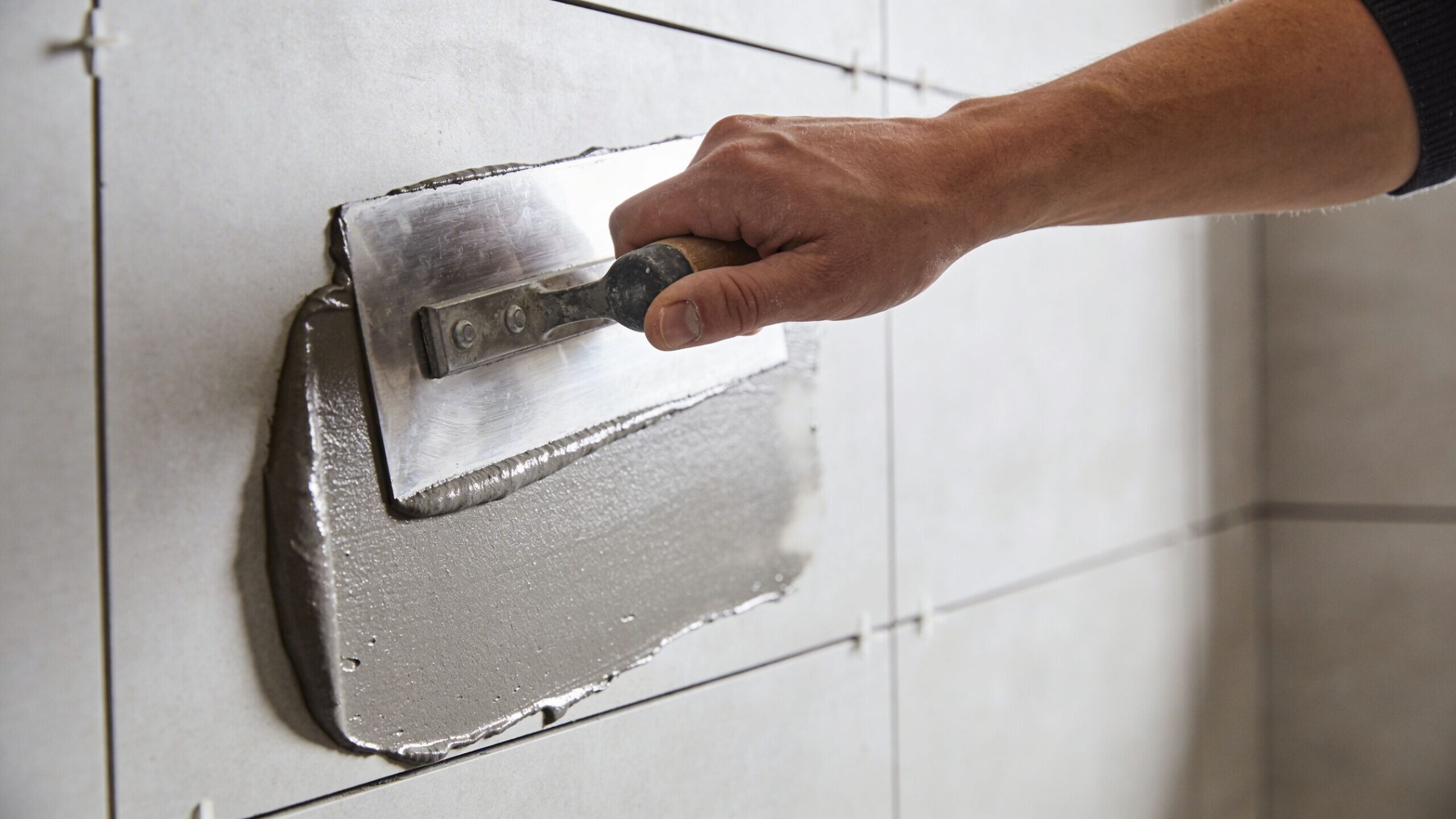 En hånd bruger en stålspartel til at påføre microcement ovenpå klinker på en flisevæg i et køkken.