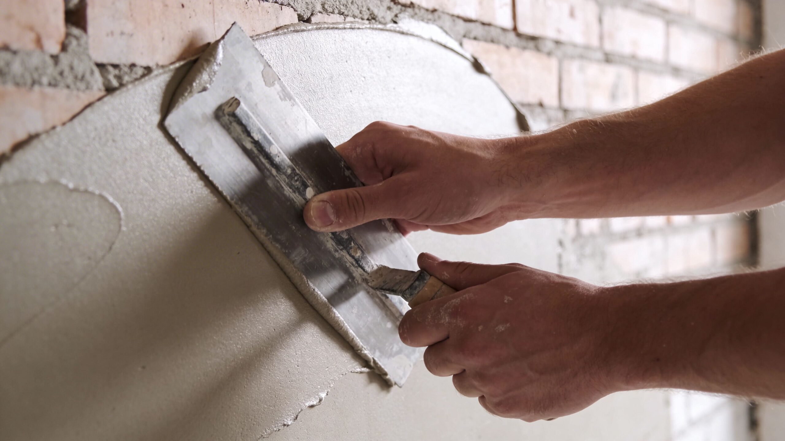En håndværker er i gang med at pudse en murstensvæg med cement og en bred stålspartel.